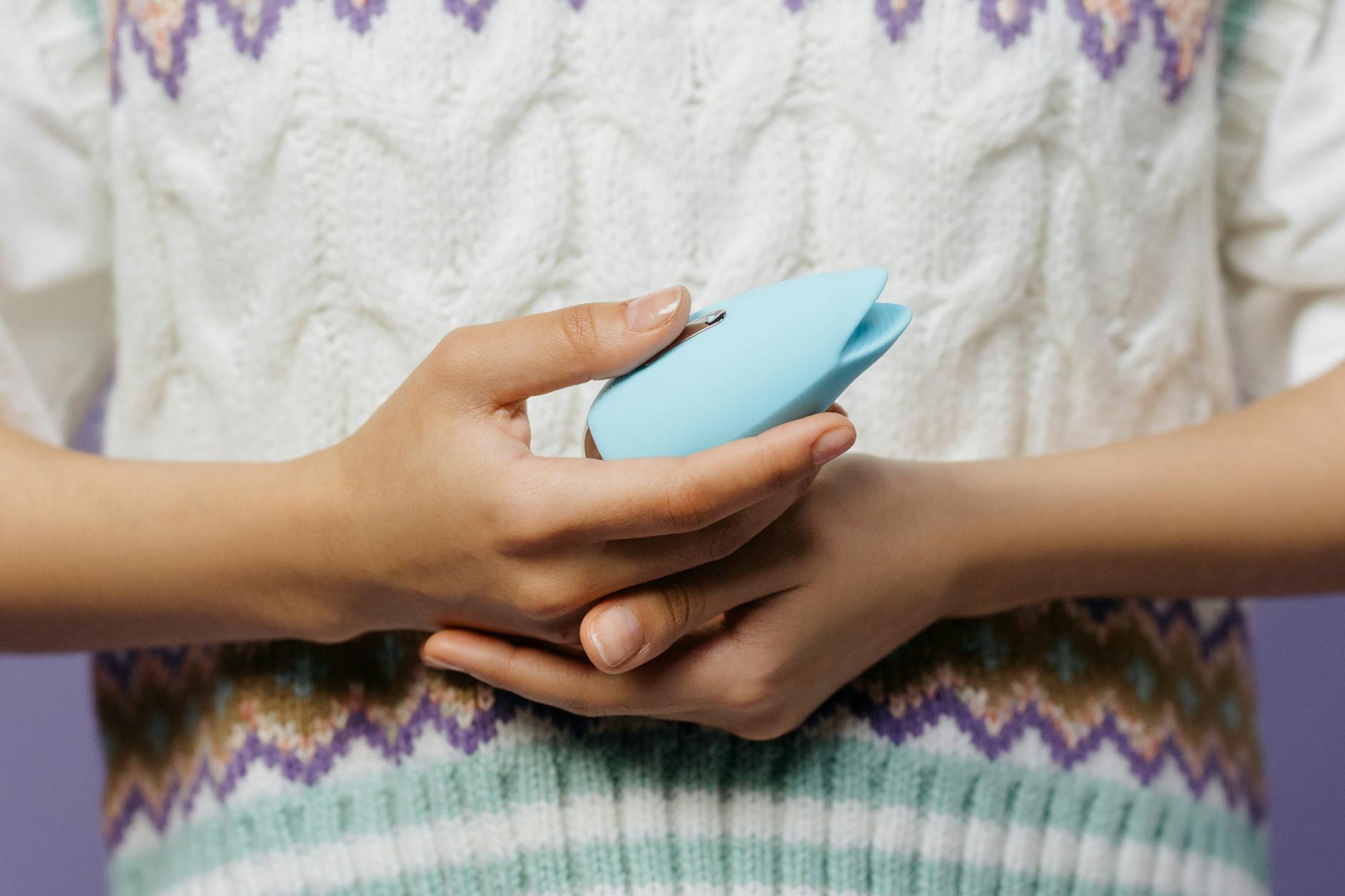 Close-up of hands holding a blue personal vibrator against a knitted sweater, representing mindful self-touch.